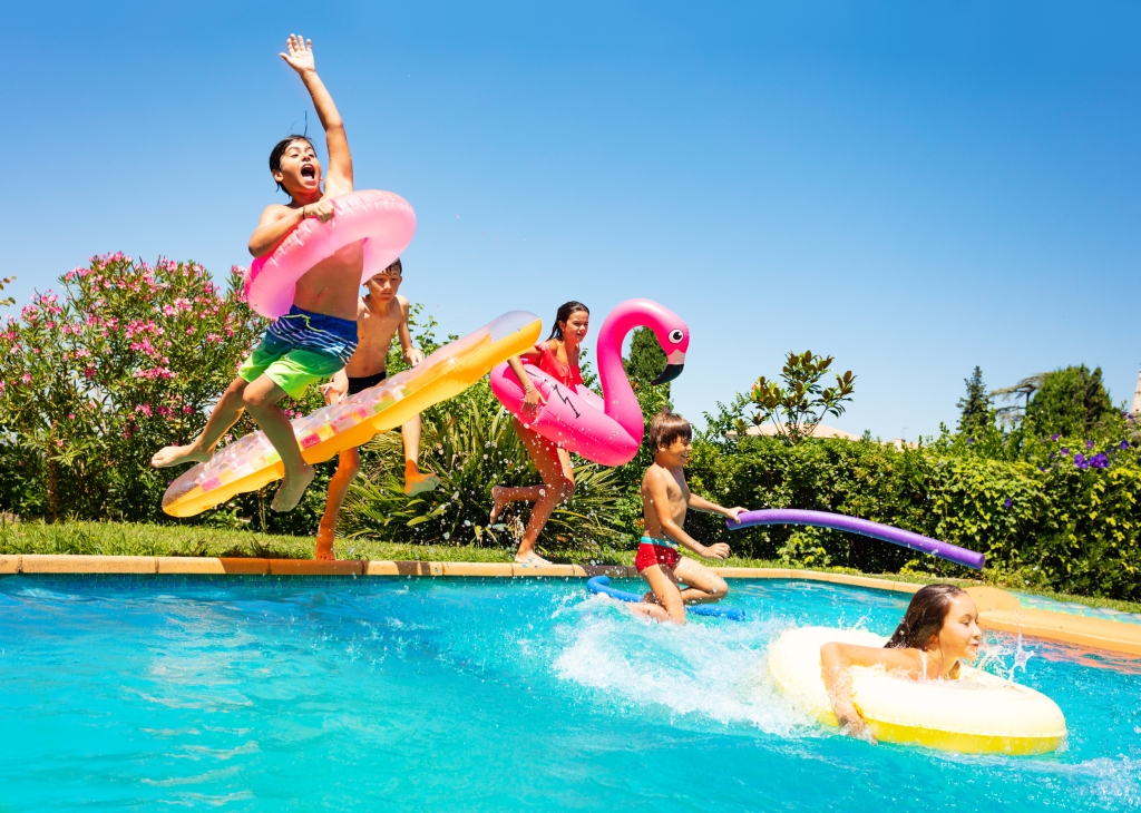 Group of age-diverse boys and girls, happy friends with swim floats jumping into swimming pool at a pool party